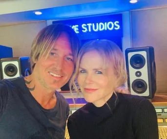 Couple smiling in a recording studio with speakers and a neon sign in the background.