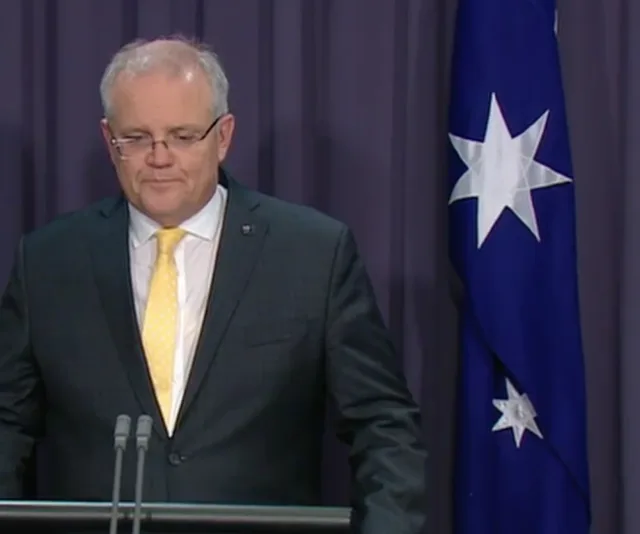 Australian official giving a press conference, standing at a podium with the national flag in the background.