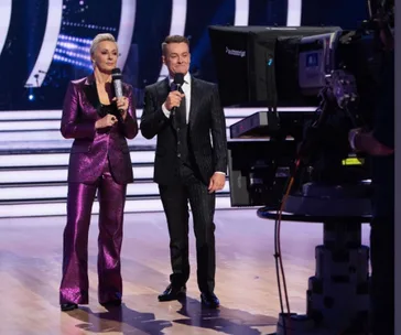 Two hosts with microphones stand on a stage during "Dancing with the Stars" 2020 finale, with cameras and lights around.