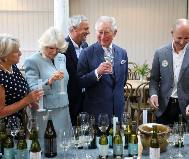 Five people smiling and tasting wine at an event, surrounded by bottles and glasses on a table.