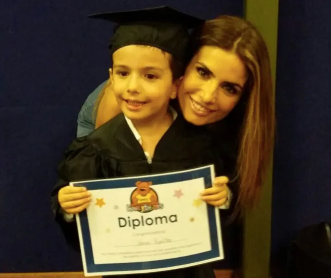 Child in graduation gown holding a diploma, smiling with a woman beside him.