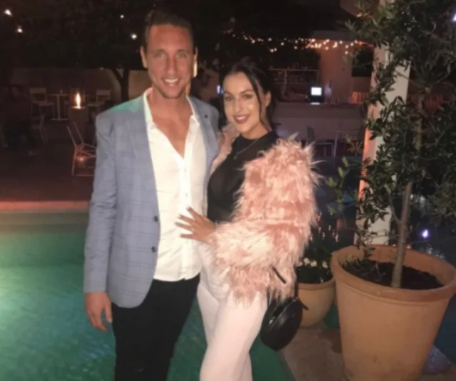 A couple poses by a pool, the man in a blue blazer, the woman in a pink fur jacket, with a potted plant nearby.