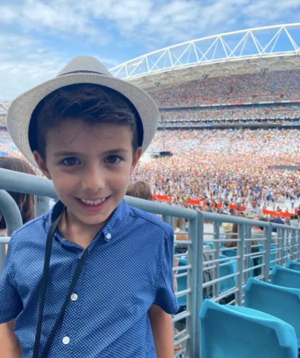 Young boy in a blue shirt and hat smiling at a crowded stadium event under a blue sky.