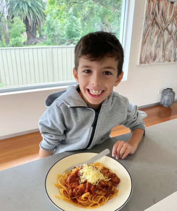 Smiling child in a grey hoodie sitting at a table with a plate of spaghetti topped with sauce and cheese.