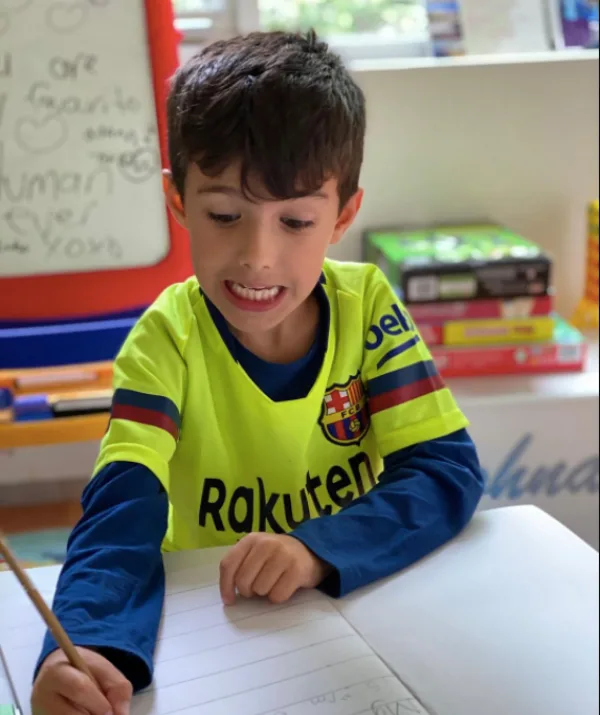 Young boy in a yellow Barcelona jersey writing in a notebook, with a playful expression, classroom background.