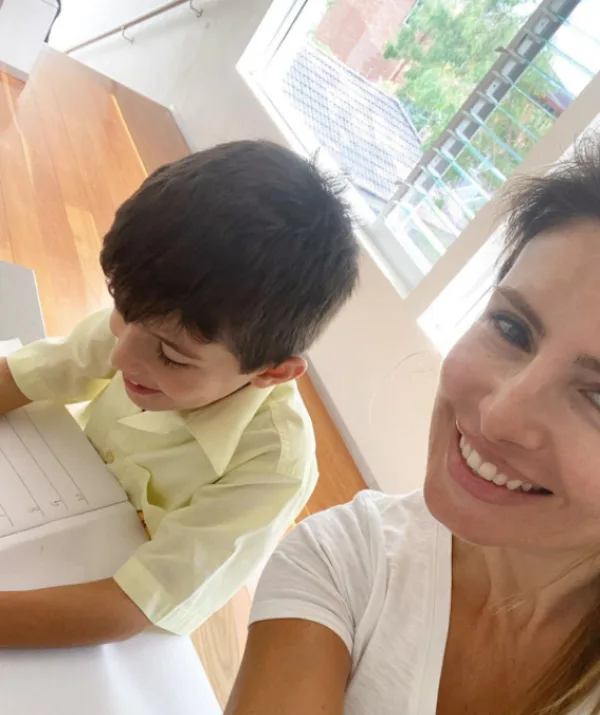 Woman smiling beside child writing in a notebook at a bright indoor space with wooden floor and window blinds.