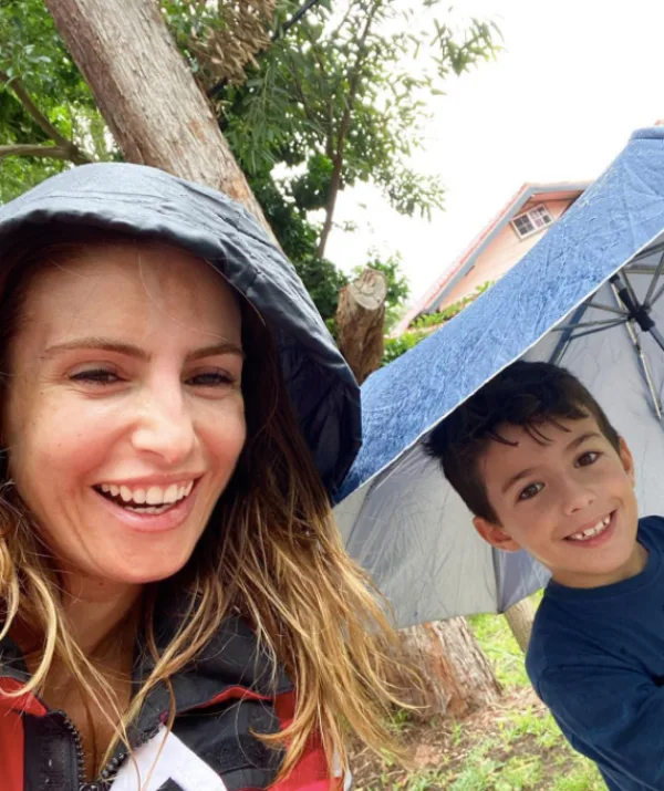 Two people outdoors; a woman smiles under a hood, and a child holds a blue umbrella, trees in the background.
