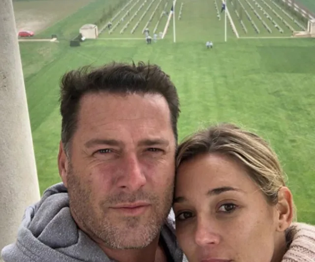 A couple takes a selfie with a vast green field and arranged chairs in the background.