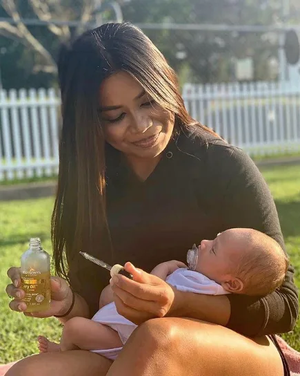 Woman sitting outdoors holds a baby, bottle of baby oil in hand, against a background of grass and white fence.