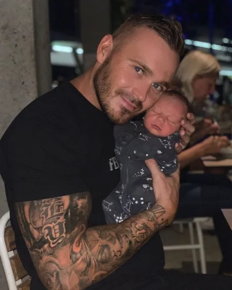 Man with tattoos lovingly holding a sleeping baby in his arms at a restaurant.
