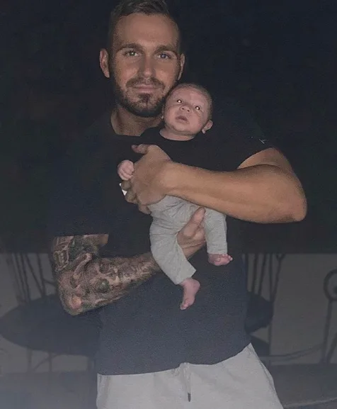 Man with tattoos holding a baby in a dimly lit setting.