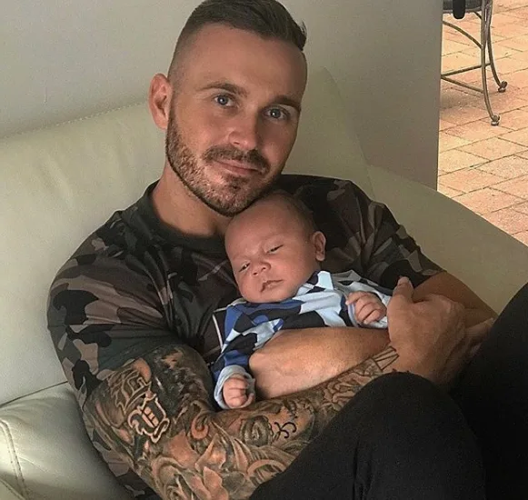 Man with tattooed arms holding a baby, sitting on a couch.