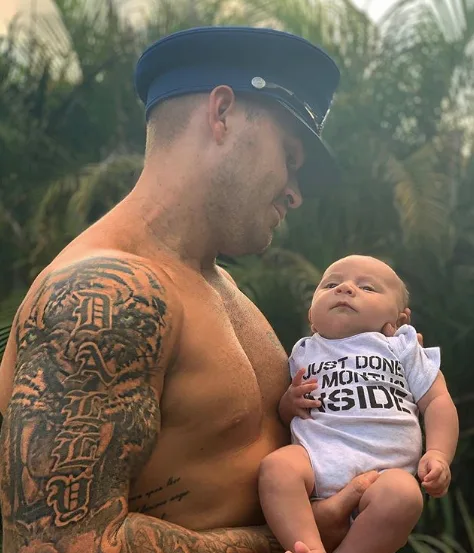 Man with tattoo and blue cap holding baby wearing "Just done 9 months inside" onesie, outdoors with palm trees.