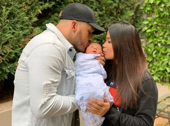 A couple kissing their swaddled newborn outside, surrounded by green foliage.