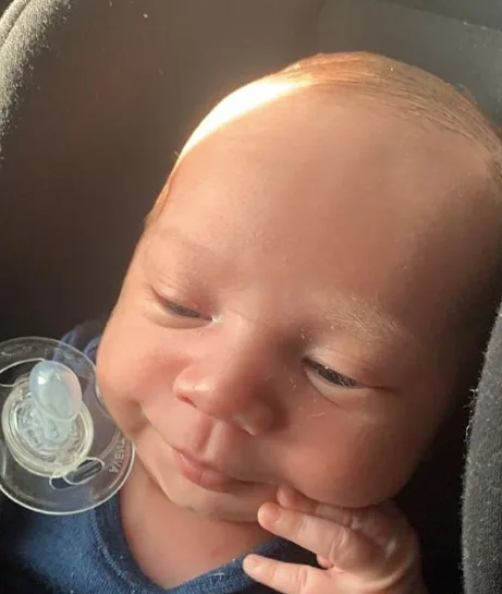 Baby with a pacifier, wearing a blue outfit, looking content in sunlight.