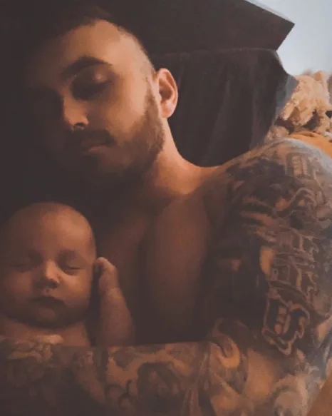 A man with tattoos cuddling a sleeping baby in his arms, both resting peacefully.