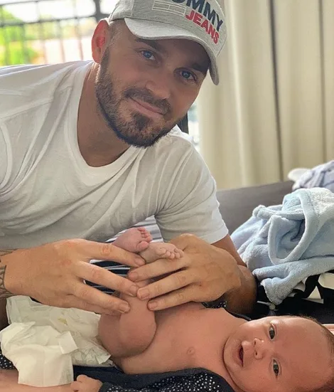 Man in a cap smiling while changing a baby's diaper at home.