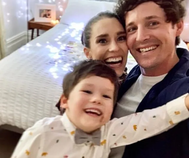 Happy family selfie in cozy bedroom with fairy lights on bed.