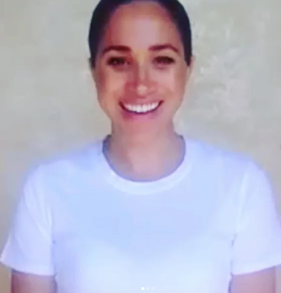 Smiling woman in a white shirt during a video call, standing against a plain background.