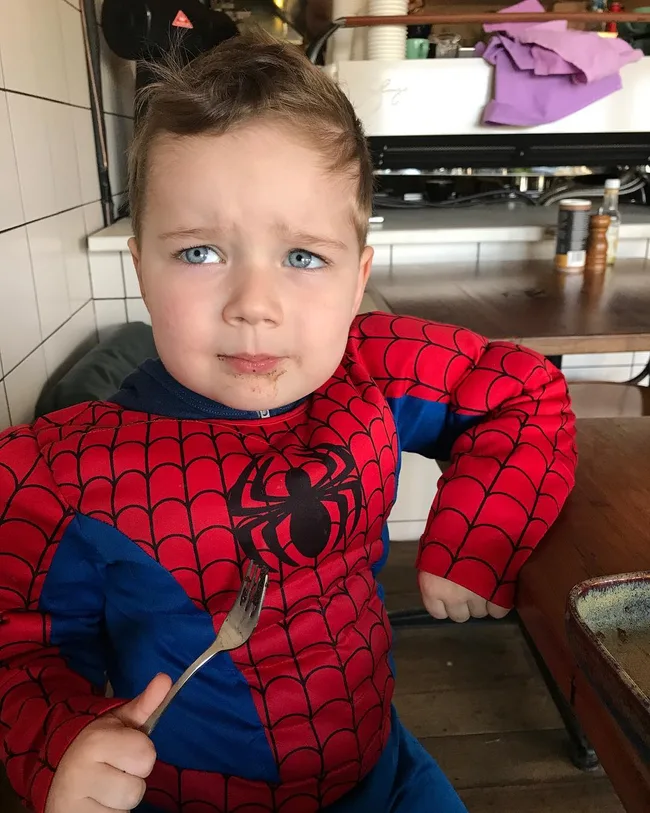 A child in a Spider-Man costume sits at a table with a fork, looking slightly puzzled.
