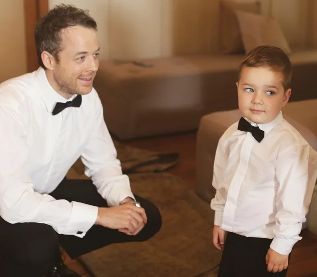 A man and a young boy wearing matching white shirts and black bow ties in a living room setting.