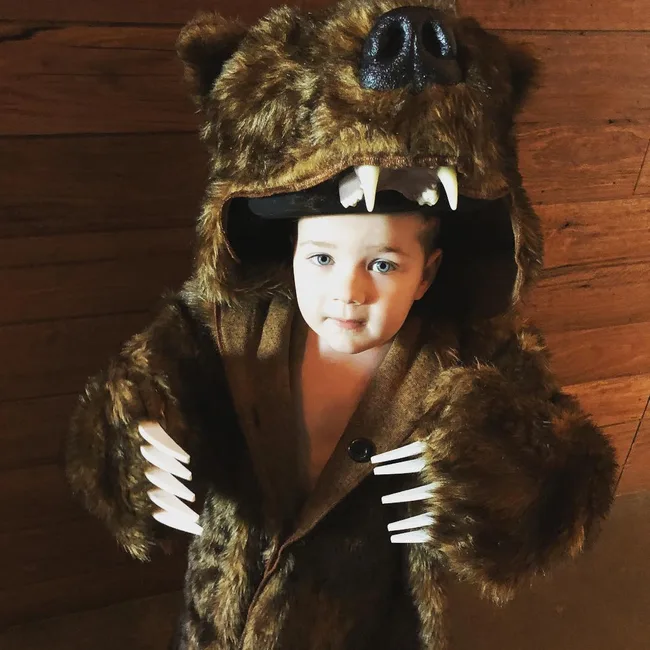 Child in a fuzzy bear costume with clawed paws and open mouth hood, standing indoors on wooden floor.