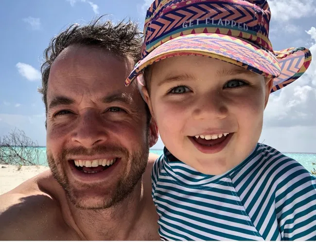 Father and child smiling at the beach, child wearing a colorful hat and striped shirt.