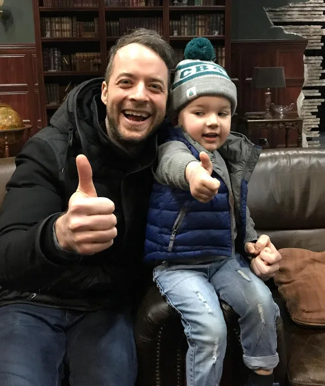 A man and a child in winter clothes giving thumbs up, sitting on a couch in a library setting.