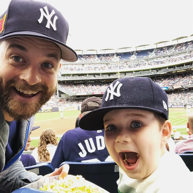 Father and child wearing Yankees caps at a baseball game, smiling with excitement, holding a tray of popcorn.