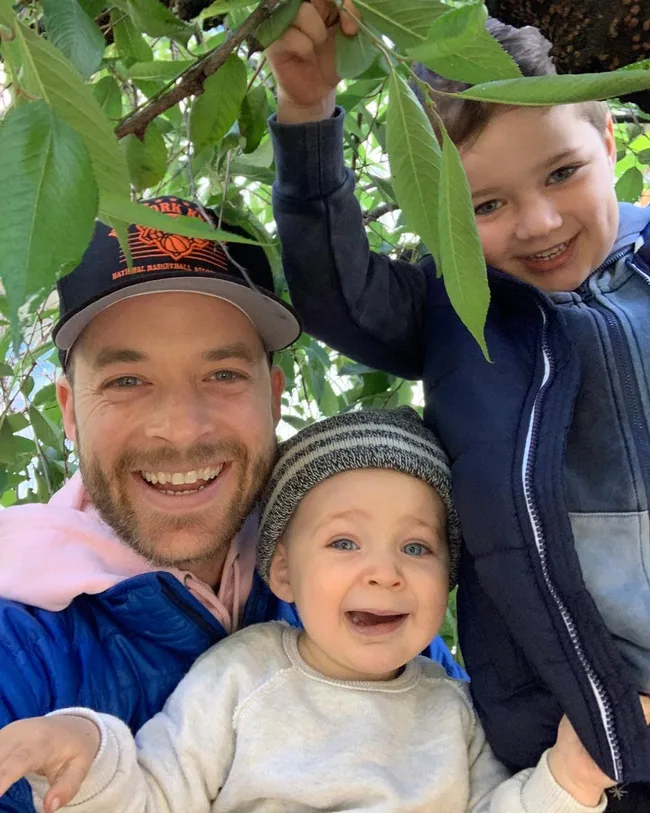 A bearded man smiles with two boys under a leafy tree; one boy is laughing, and the other is wearing a beanie, looking surprised.
