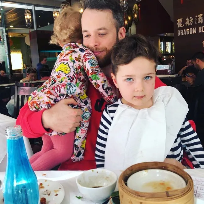 A man holding a toddler with a child next to them at a restaurant table with food and a blue bottle.