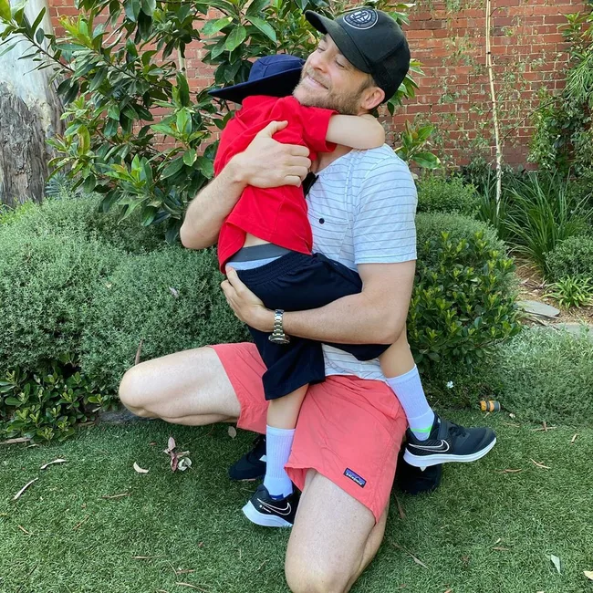 Man in cap kneeling and hugging a child in red shirt and navy shorts, set in a garden with green shrubs.