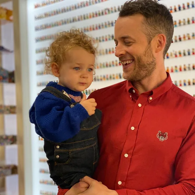 Man in red shirt holding a child in a blue sweater, smiling in a room with small figurines on the wall.