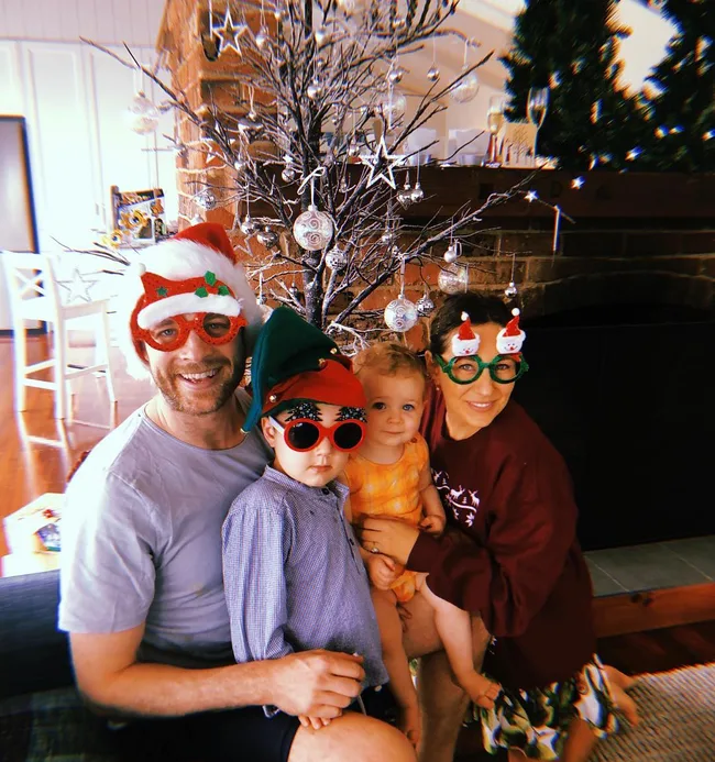 Family in festive glasses and hats pose with two children in front of a decorated tree indoors.