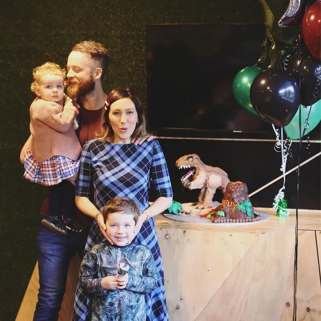A family with two kids at a party with dinosaur-themed cake and balloons.