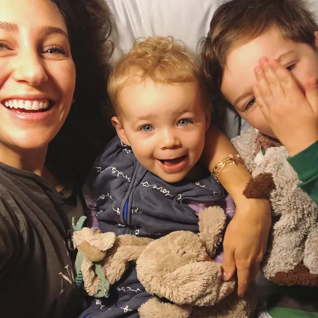 A woman with two children, smiling and cuddling on a bed with soft toys.