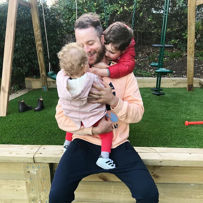 Man sitting on grass, holding two young children in a backyard with a swing set in the background.