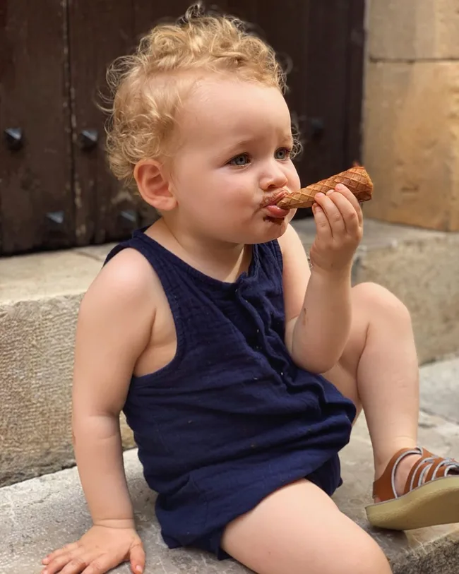 Young child with curly hair, wearing a navy outfit, enjoying an ice cream cone while sitting on stone steps.