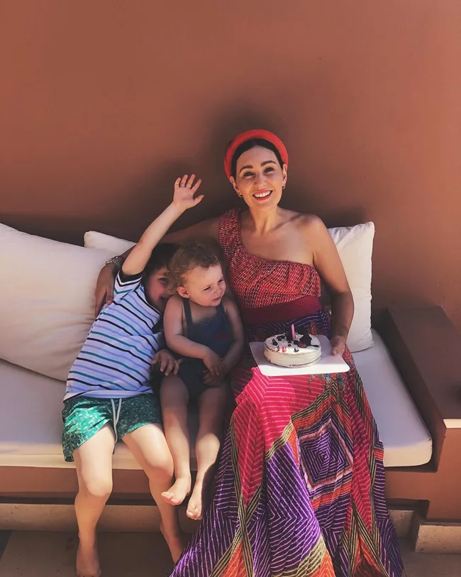 Woman in colorful dress holds cake, sitting with two children on a couch outdoors.