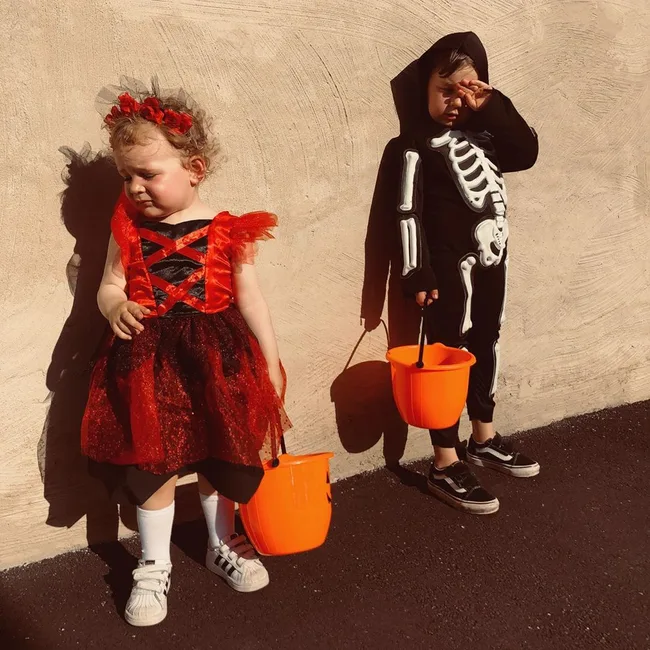 Two children in Halloween costumes, a skeleton and a witch, holding orange buckets against a textured wall in sunlight.