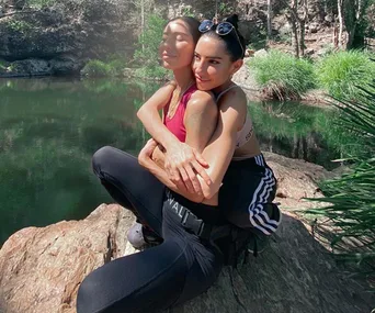 Two women sit embracing on a rock by a serene forest lake, with greenery in the background.
