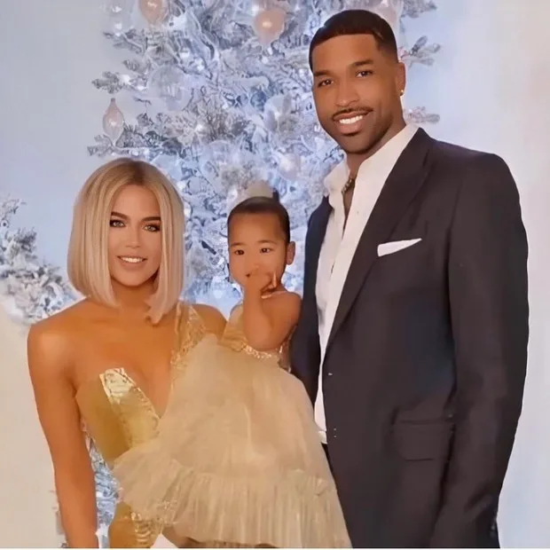 A family posing in front of a decorated Christmas tree; the woman in gold, child in cream, and man in a black suit.