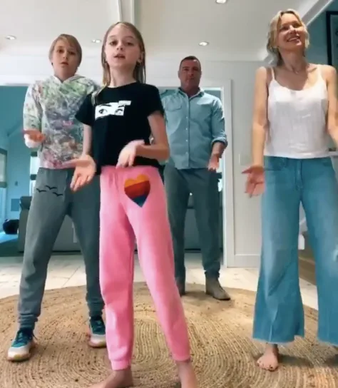 A family of four dancing happily together in a well-lit living room, wearing casual, colorful clothing.