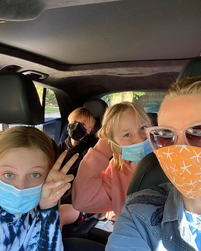 Group of four people in a car wearing colorful face masks and sunglasses, smiling and making peace signs.