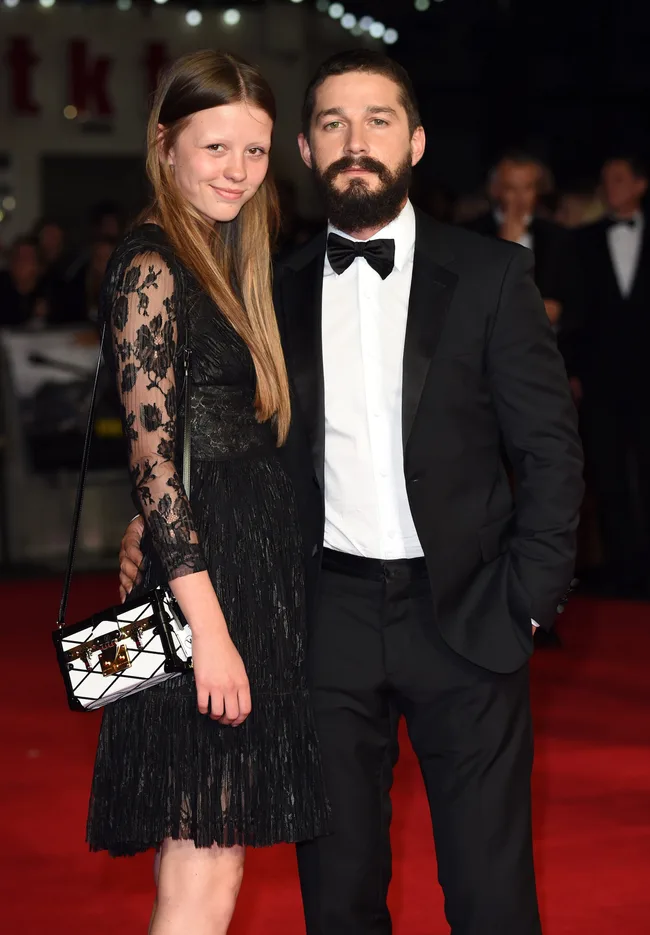 A couple poses on a red carpet; the woman in a black lace dress and the man in a black suit with a bow tie.