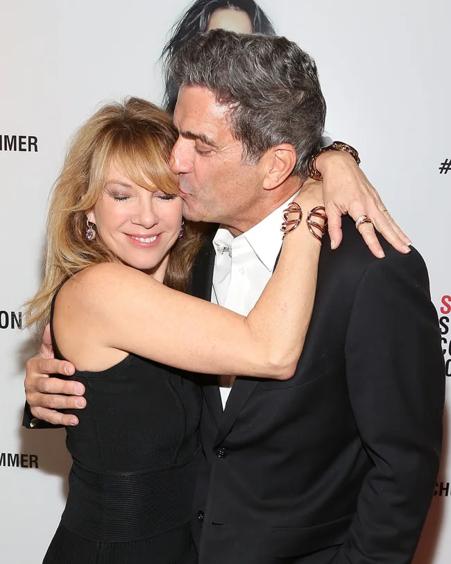 Couple embracing; man in suit kisses woman's cheek at an event with a poster in the background.
