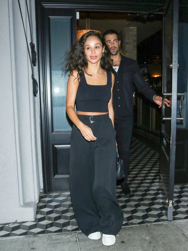 A woman in a black outfit and white sneakers exits a door, followed by a man in dark clothing.