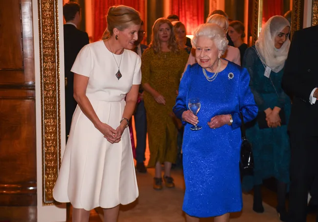 Two elegantly dressed women conversing at a formal event, one in blue holding a drink, surrounded by guests.