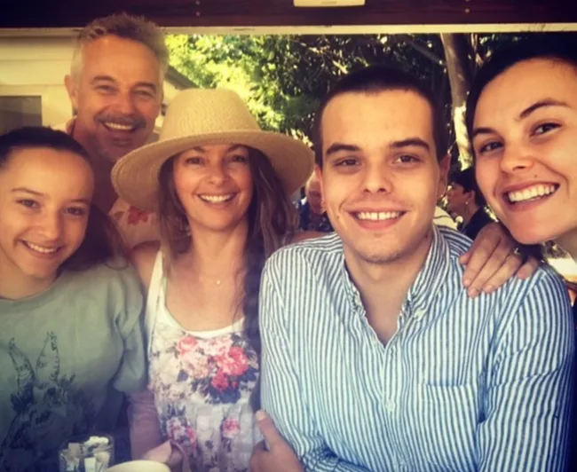 A smiling family of five posing outdoors, with one wearing a straw hat.