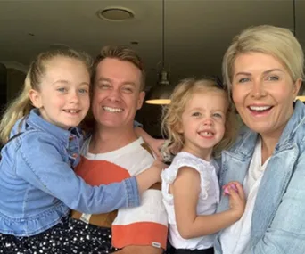 A family of four smiling indoors, with two young girls held by their parents.